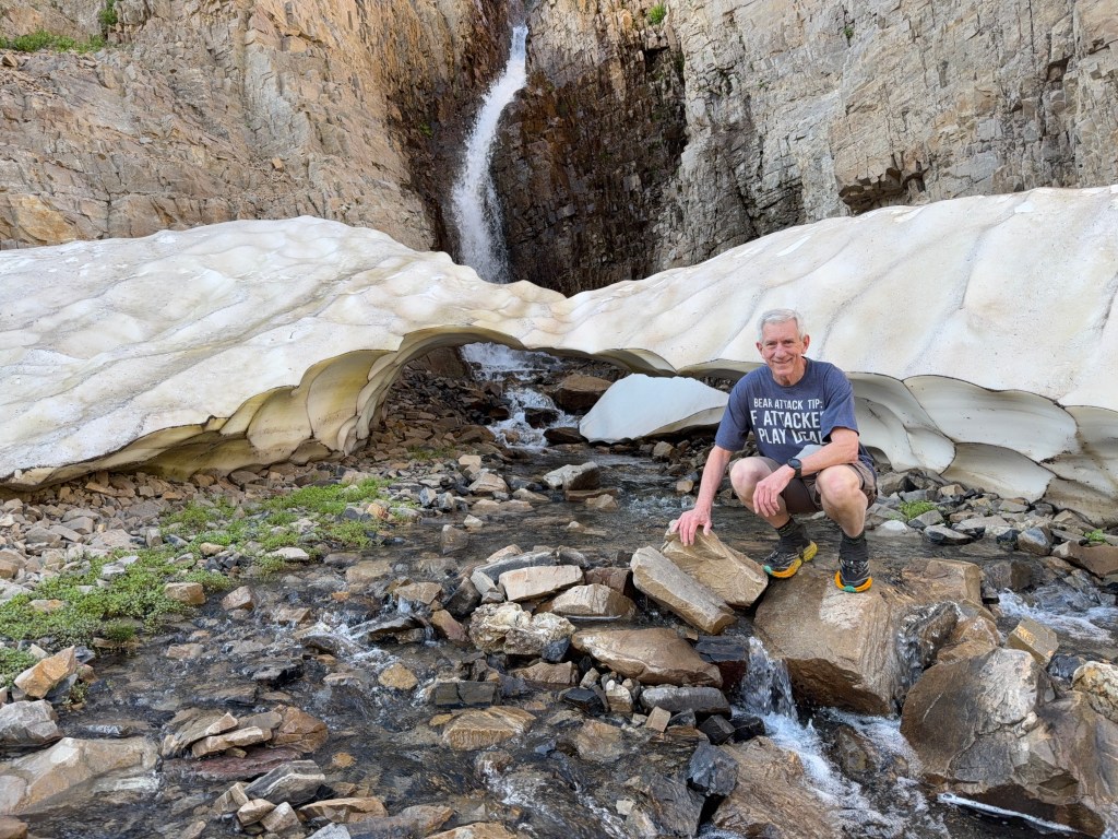 Old Hiking Dude kneeling by a creek, snow bridge and waterfall